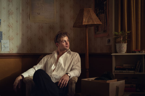 Leo Woodall as Dexter in One Day, sitting in a living room wearing a white shirt and black trousers.