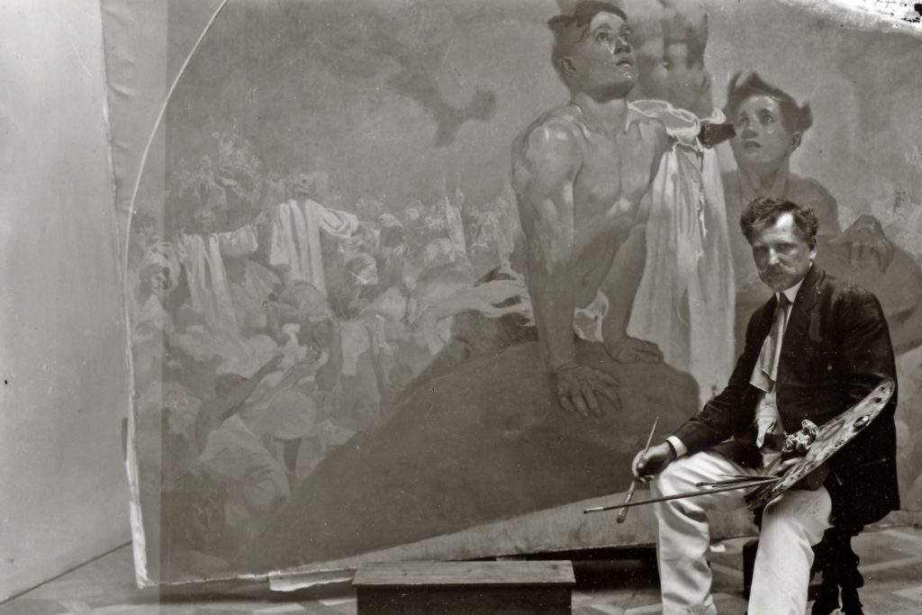 Black and white image of Alphonse Mucha completing a painting.