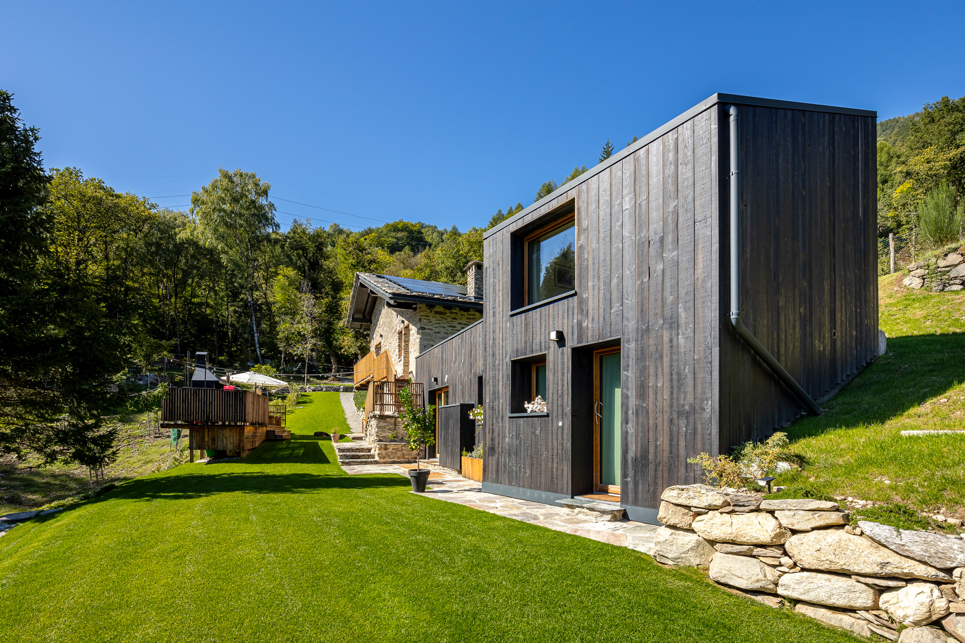 Lodge clad in black wooden panels, with a stone path and views of the Italian hills. 