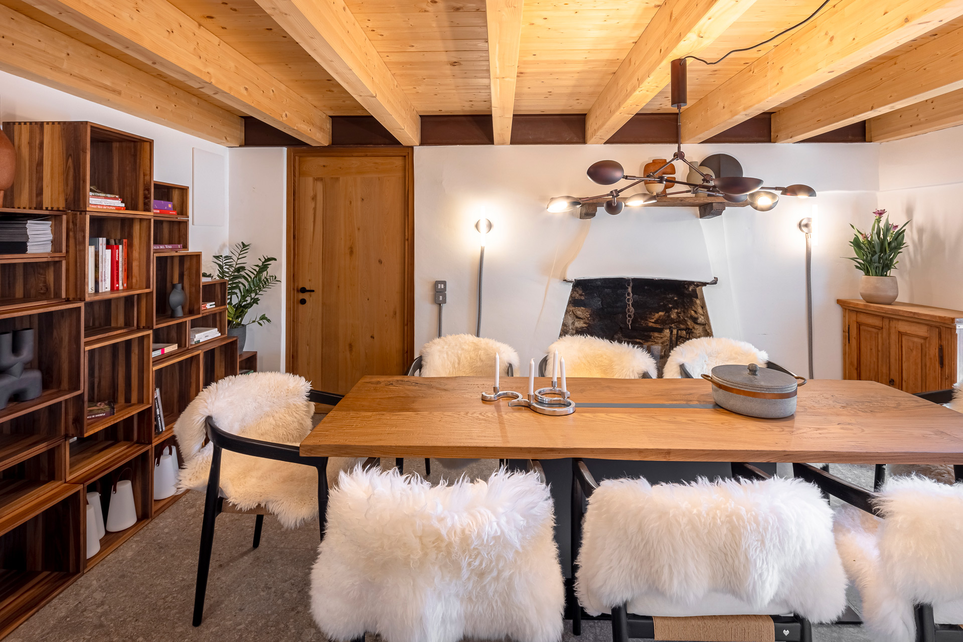 Dining room with wooden table, bookshelves and ceiling beams, with fur-covered chairs.