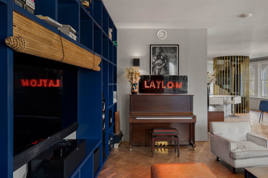 Living room with navy walls, large TV, parquet flooring and a piano with a Laylow neon sign above