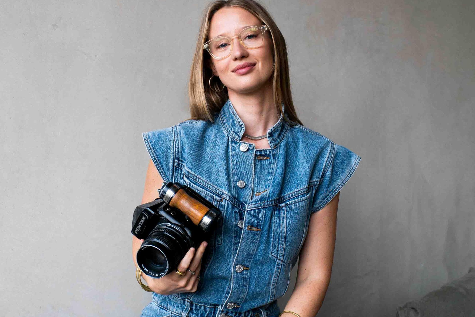 Alice Aedy wearing a cap-sleeve denim jumpsuit and holding a camera.