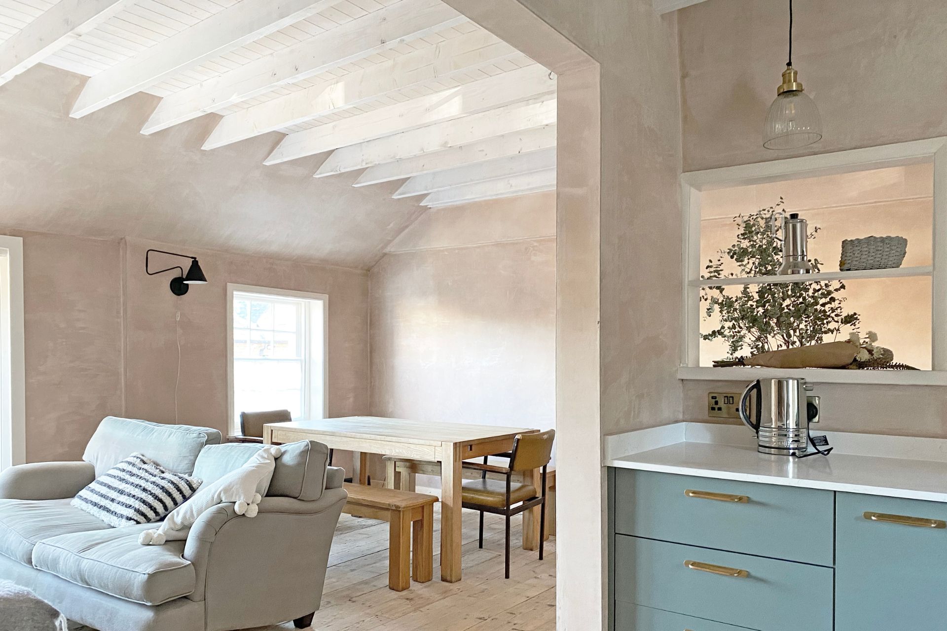 Living room with washed terracotta walls, wooden furniture, a grey sofa and blue kitchen cabinets.