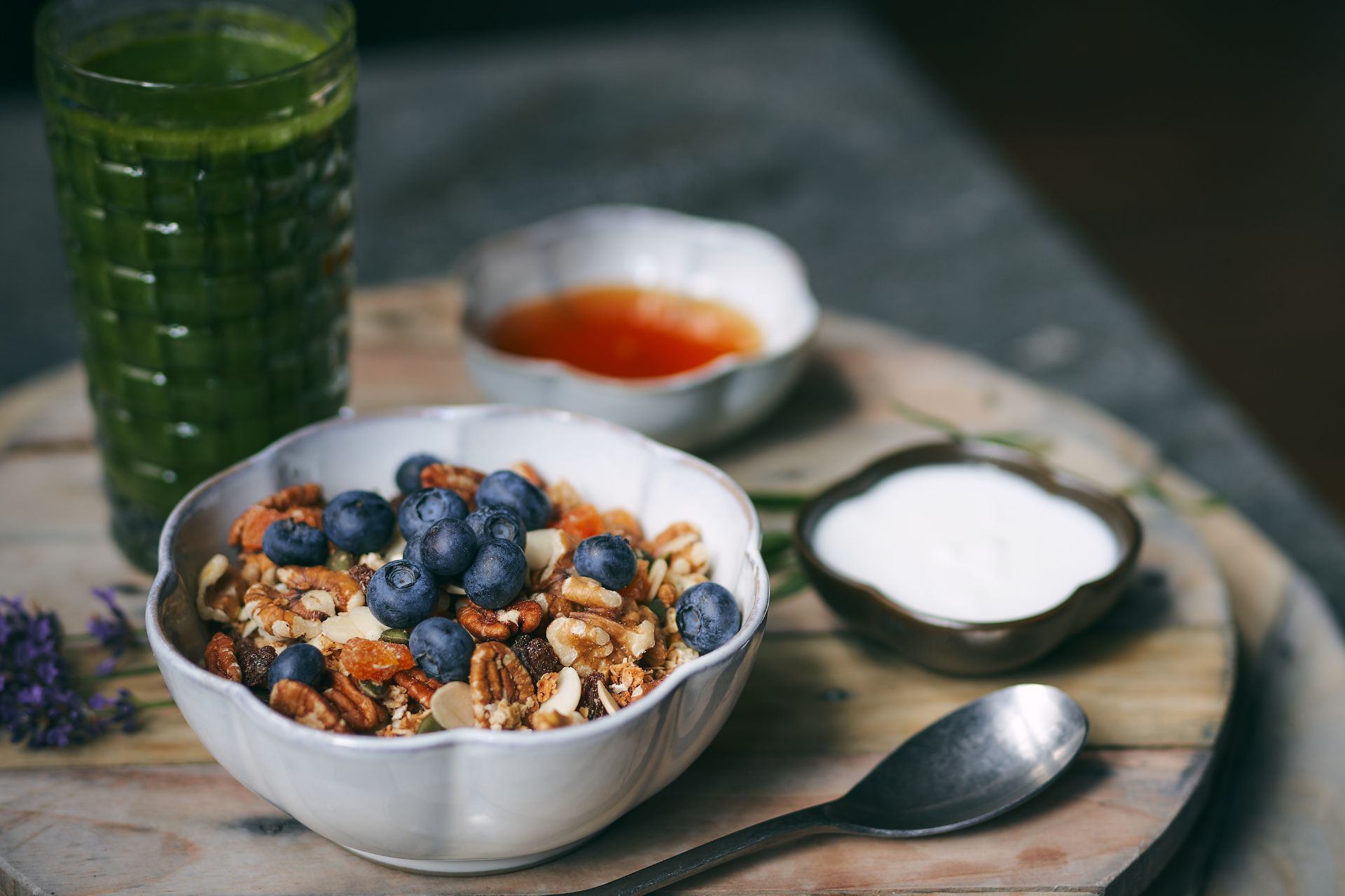 White ceramic bowl of granola and blueberries, with a glass of green juice beside it.