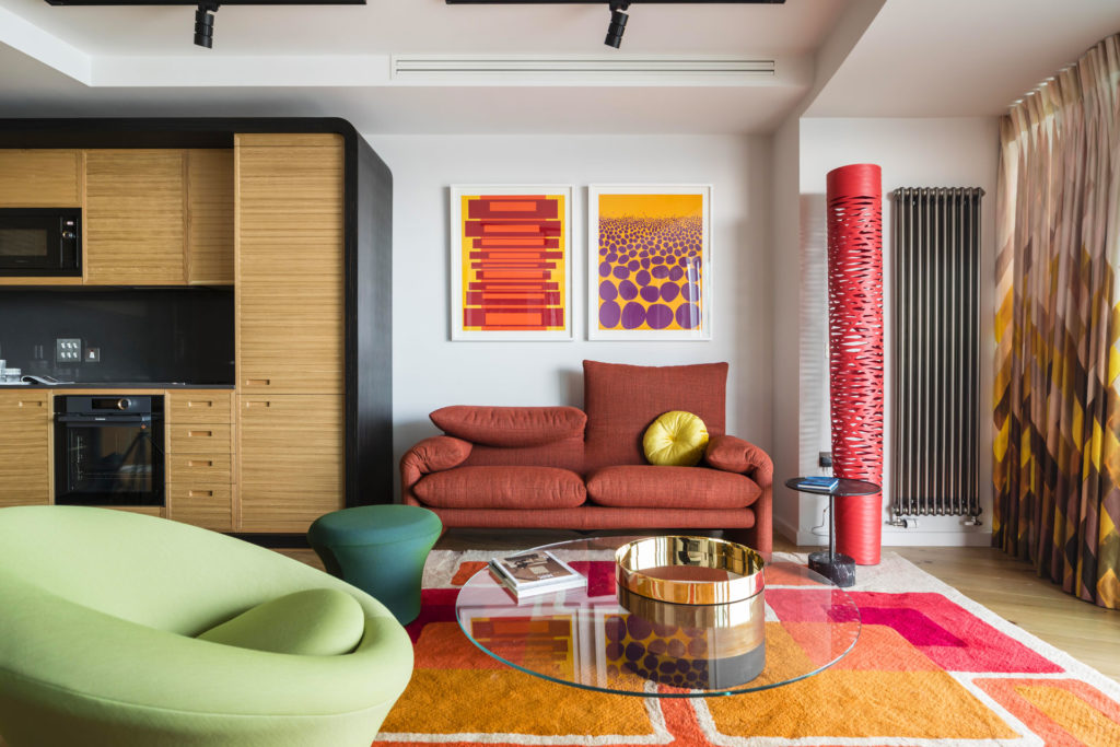 A brightly coloured, 70s-inspired living room. There is a red sofa in the centre with art behind it.