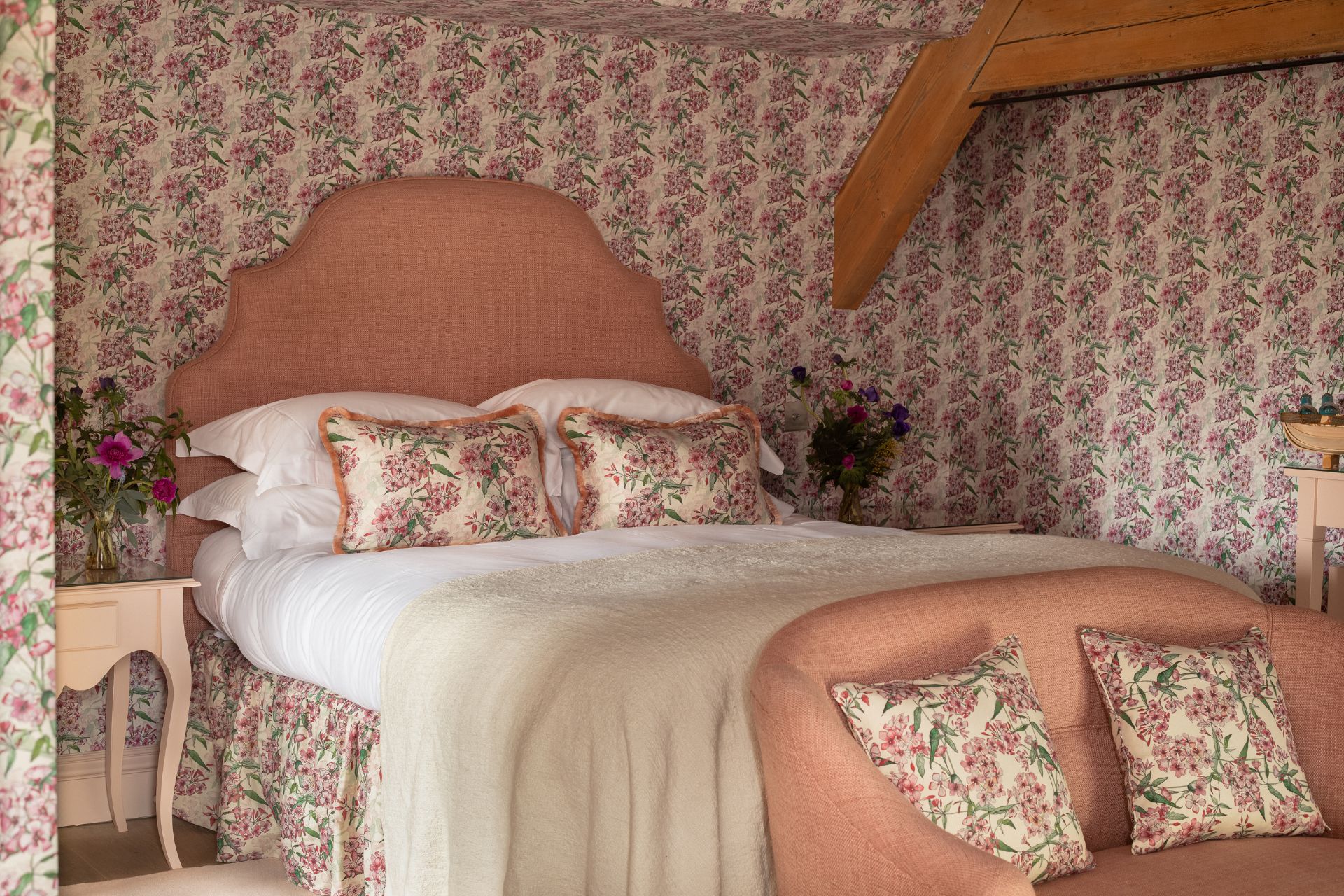 Hotel room with pink and white floral wallpaper, s pink headboard on the bed, and white bed linen.