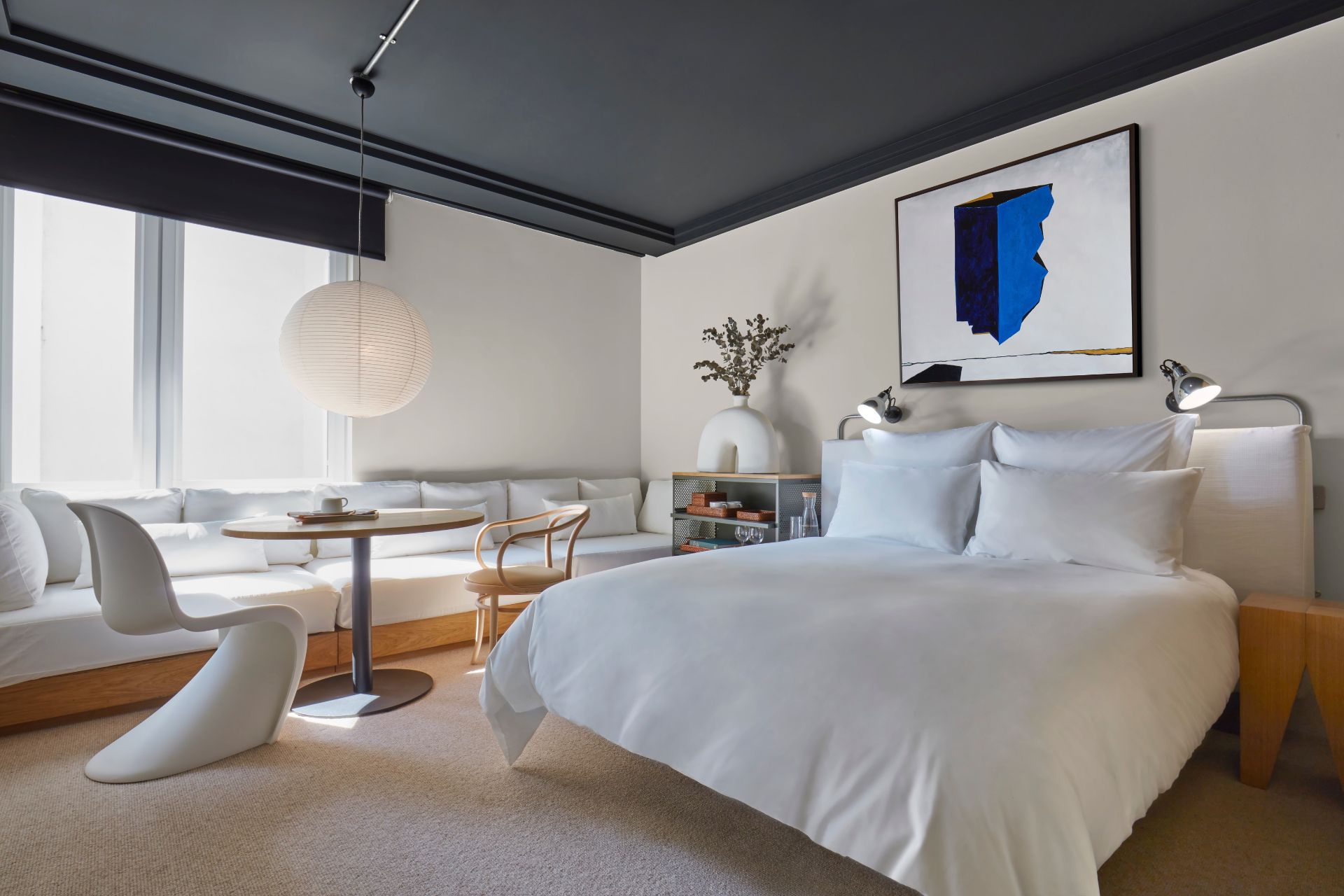 Bedroom with white bed linen, a white chair and paper lampshade, and a blue abstract print above the bed.