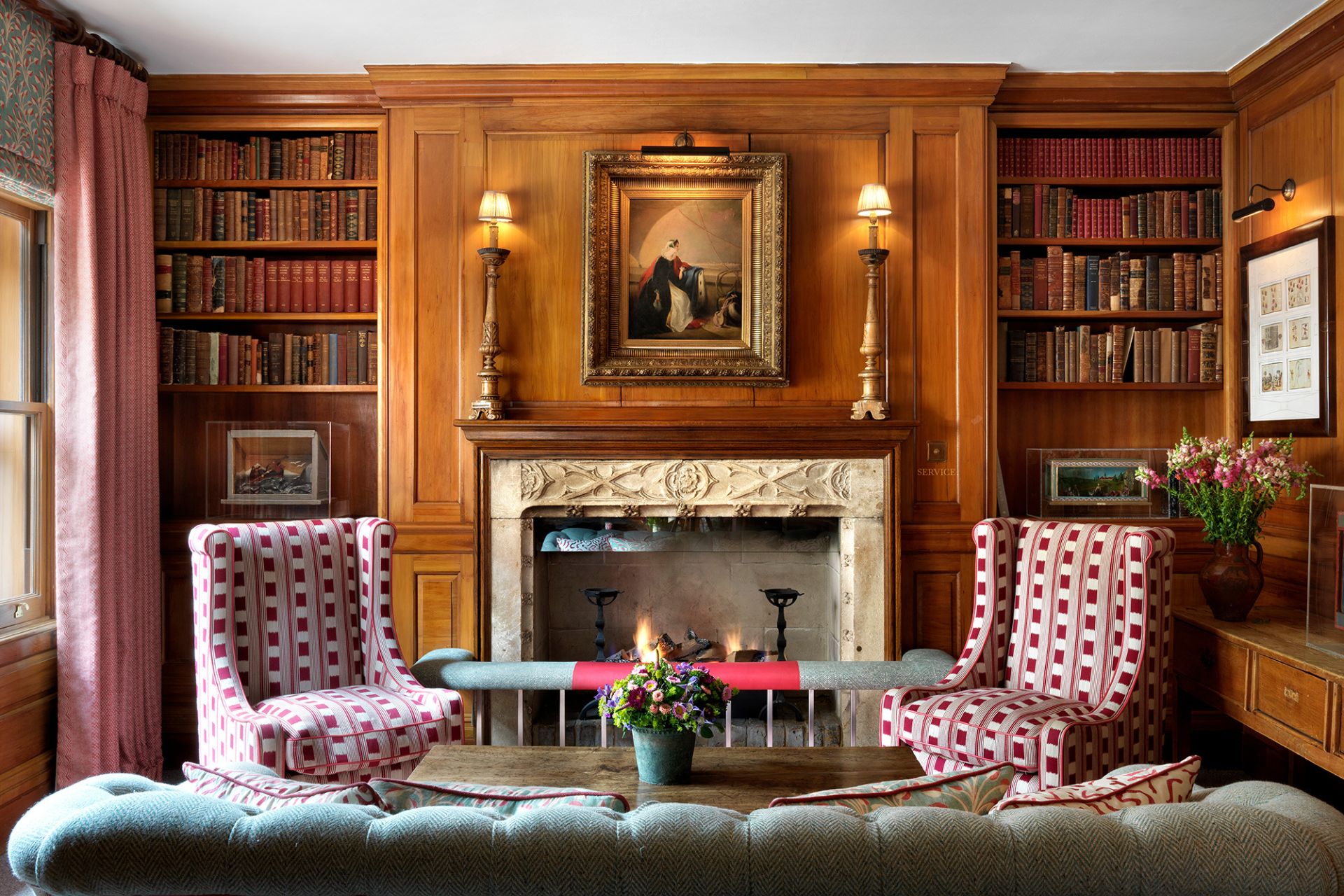 Wood-panelled library with red armchairs and a central fireplace.