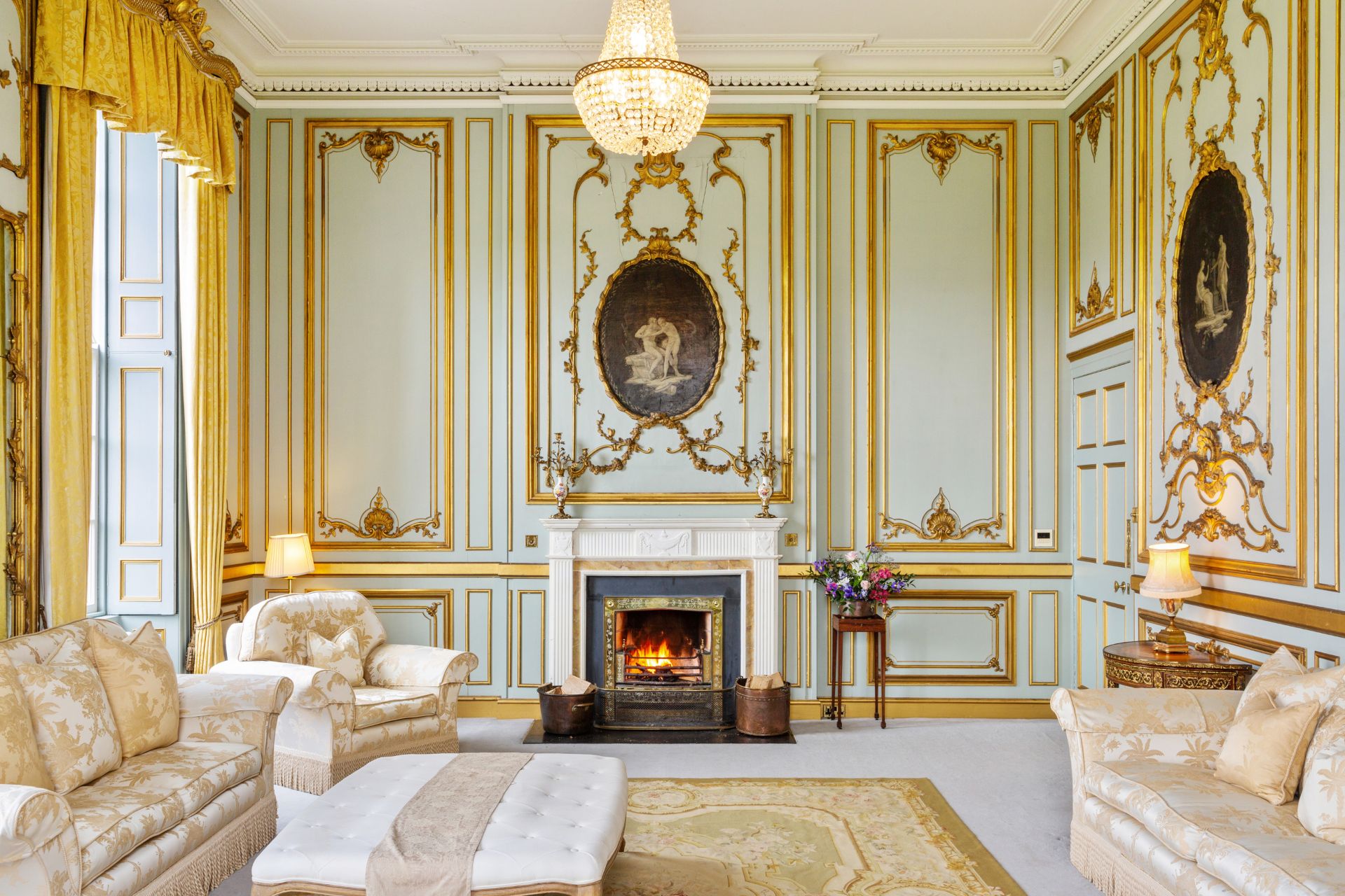 Living room with cream and gold sofas, yellow drapes a chandelier, and gilt detailing on the walls.