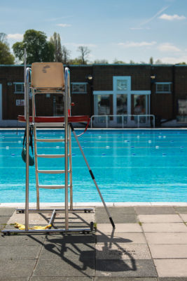 A Lido - outside swimming pool with lifeguard chair in foreground - in the distance the pool glistening in the sunshine, the lane markers are clear and the depths of water are just visible.