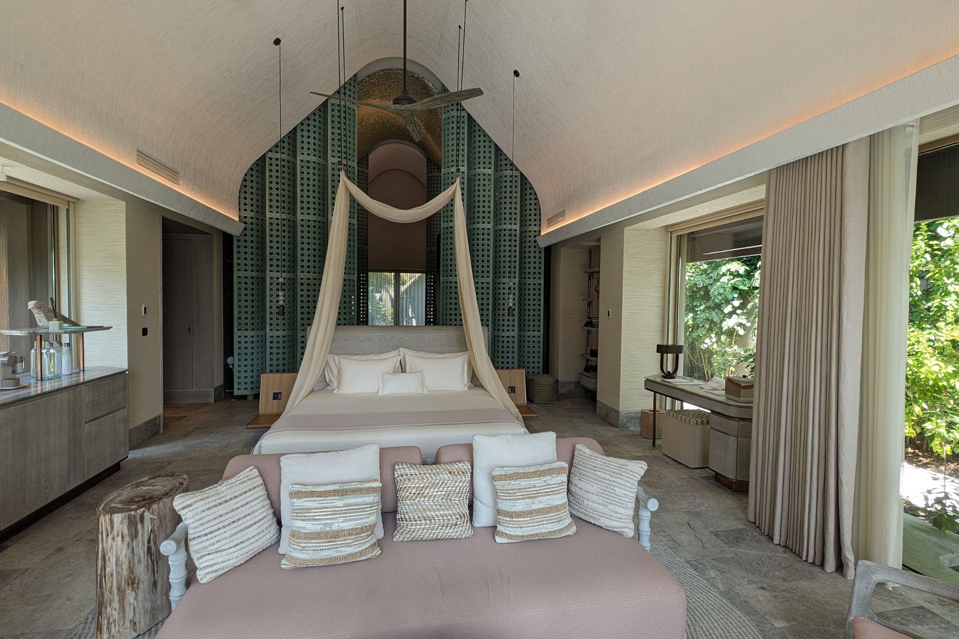 Bedroom with canopy bed, pink chaise longue and a view of palm trees outside.