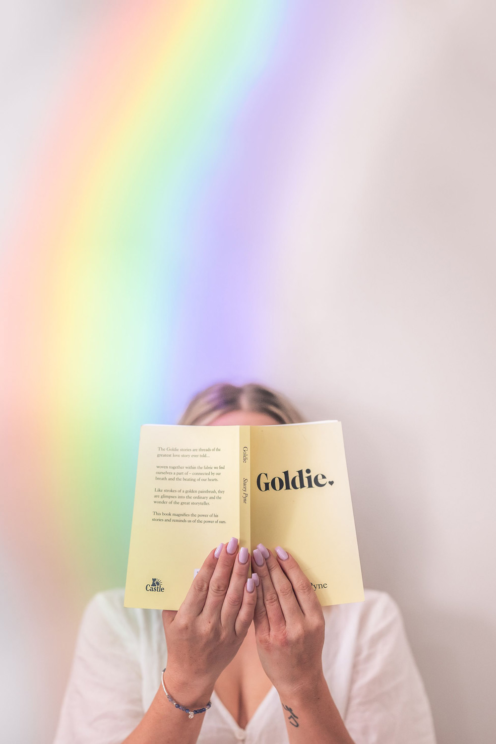 A woman with a book in front of her face and light casting a rainbow over her and the white wall behind her