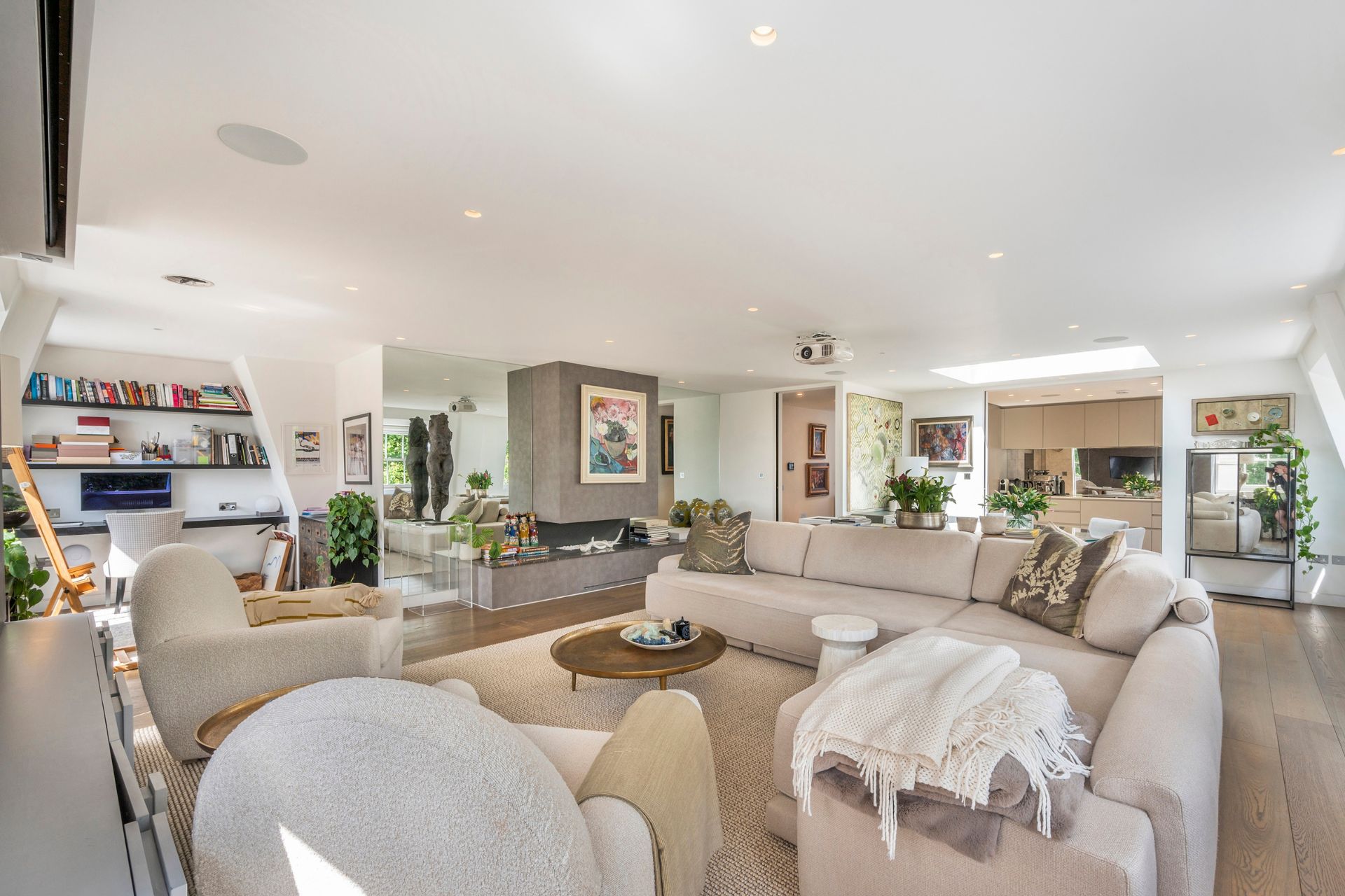 Living room with cream sofas, bookshelves and view of a modern kitchen with wooden cabinets.