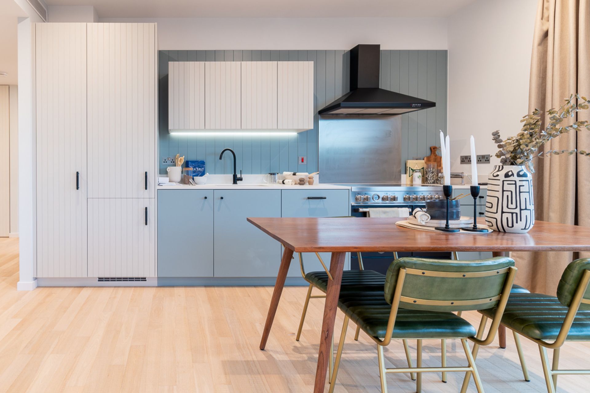 Kitchen with pale blue cabinets, a black extractor fan and a wooden dining table with forest green velvet chairs