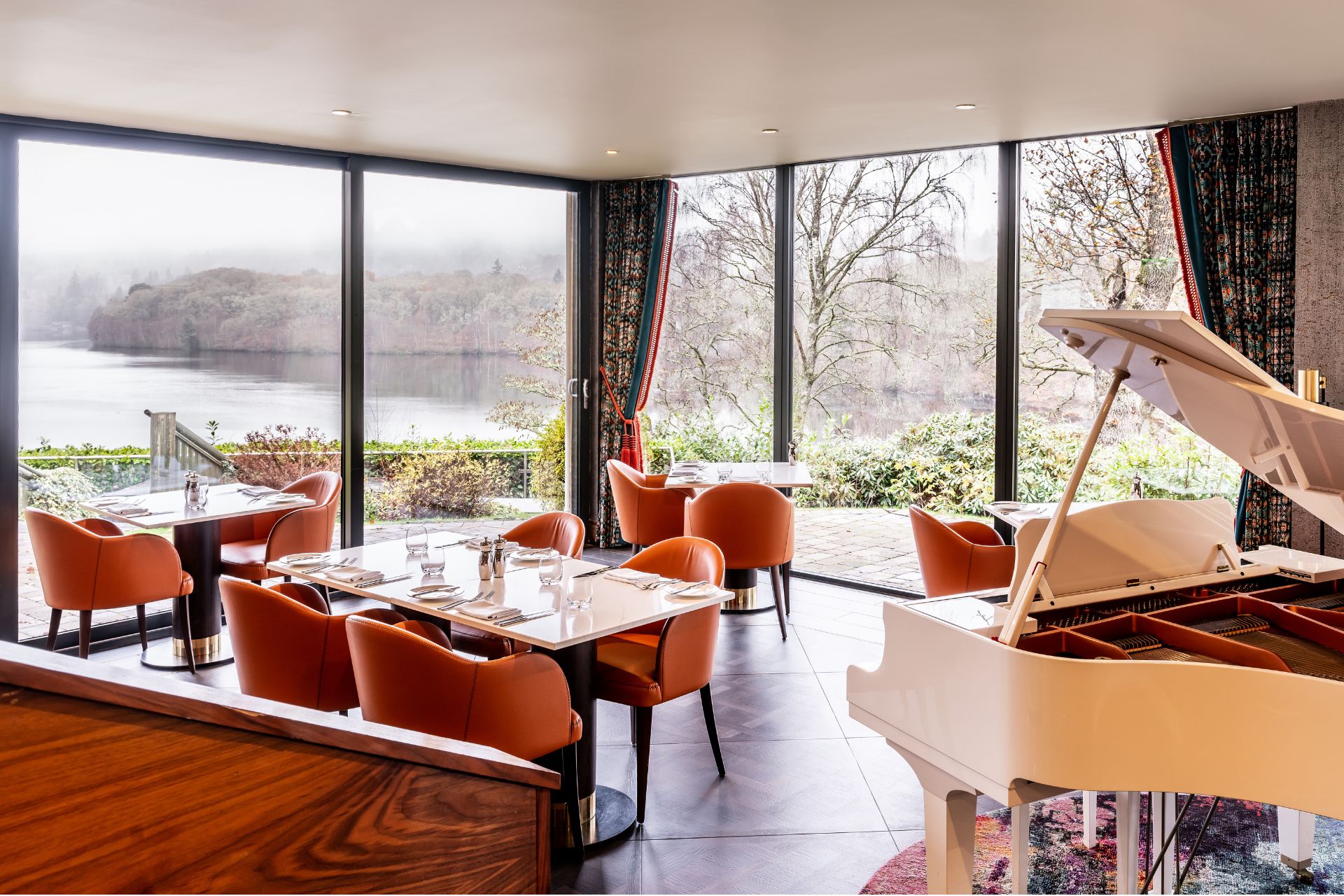 Restaurant with orange armchairs, white grand piano and floor-to-ceiling windows overlooking the Scottish hills
