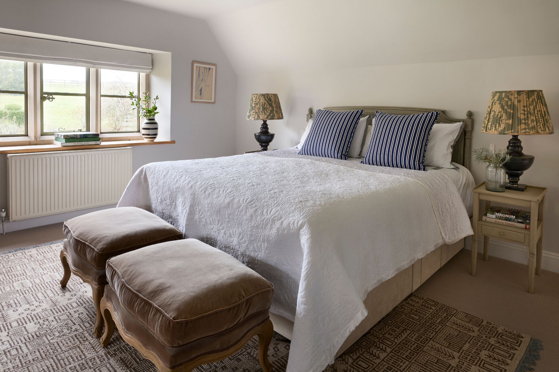 Bedroom with white bedspread, blue striped cushions and two brown velvet stools.