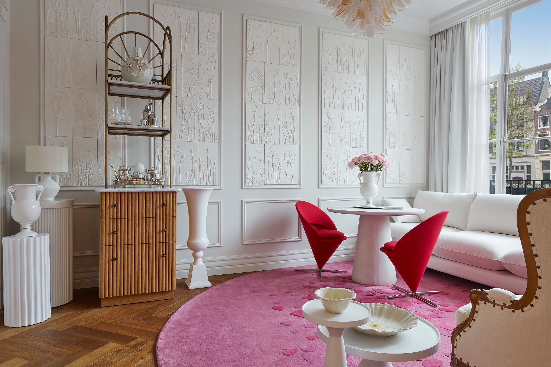 White hotel suite with pink rug, red chairs and a wooden bar cart.