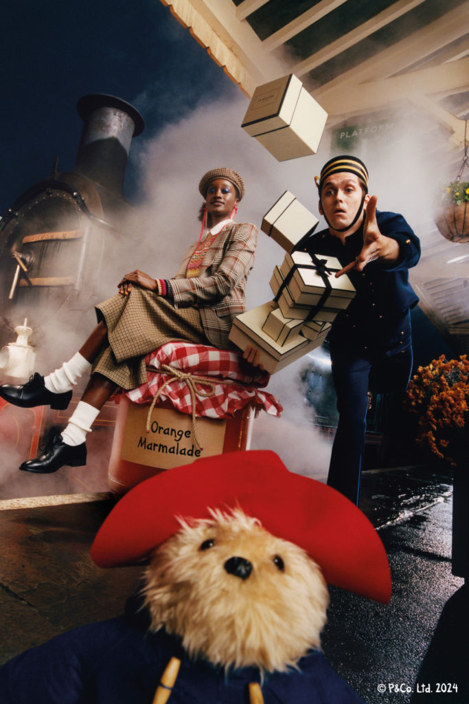 Woman sat on luggage on train station platform next to station staff dropping boxes with teddy bear in foreground