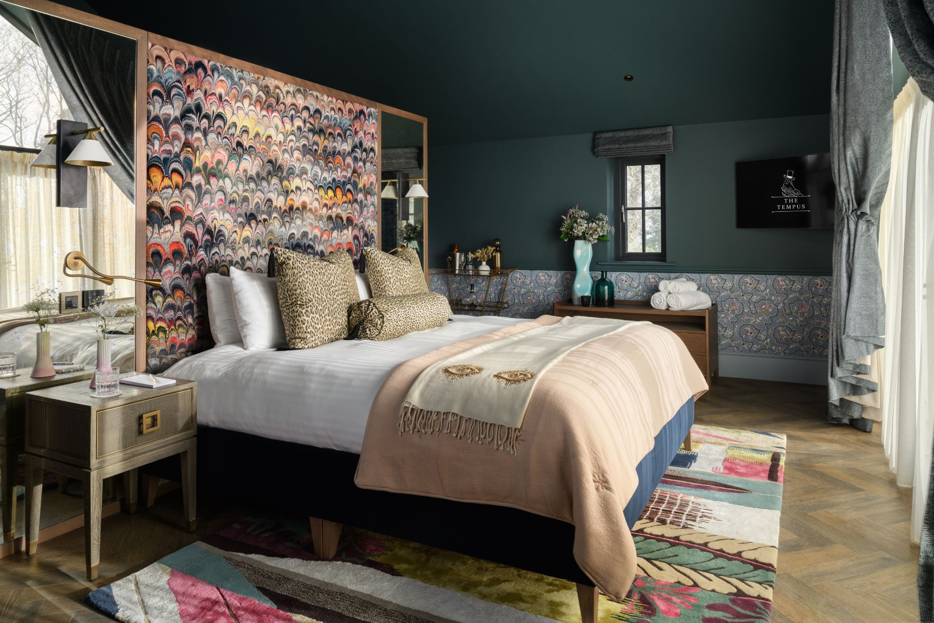 Hotel bedroom with patterned headboard, beige bedspread and leopard print cushions.