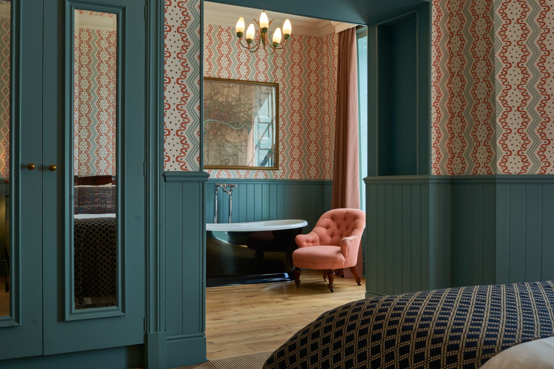 Hotel bedroom with teal panelling, pink floral wallpaper and a black freestanding bathtub with a pink velvet chair beside it.