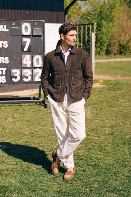Man stood on cricket ground wearing white trousers and bronw jacket
