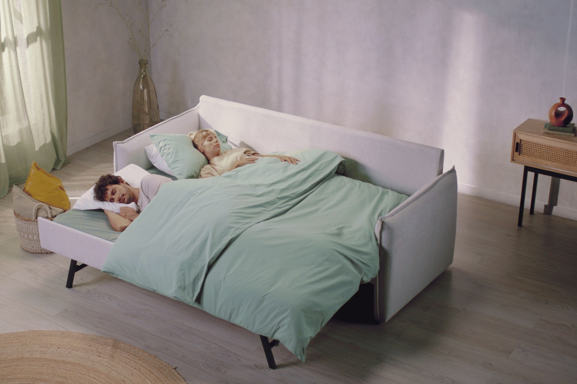 White sofa bed with two people sleeping on it under a green blanket