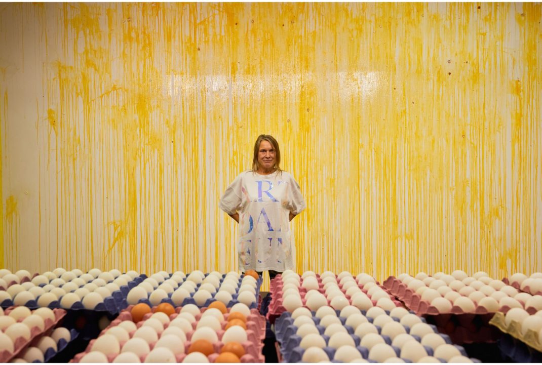 Artist Sarah Lucas stands in front of wall covered in broken eggs, with rows of unbroken eggs in boxes in front of her