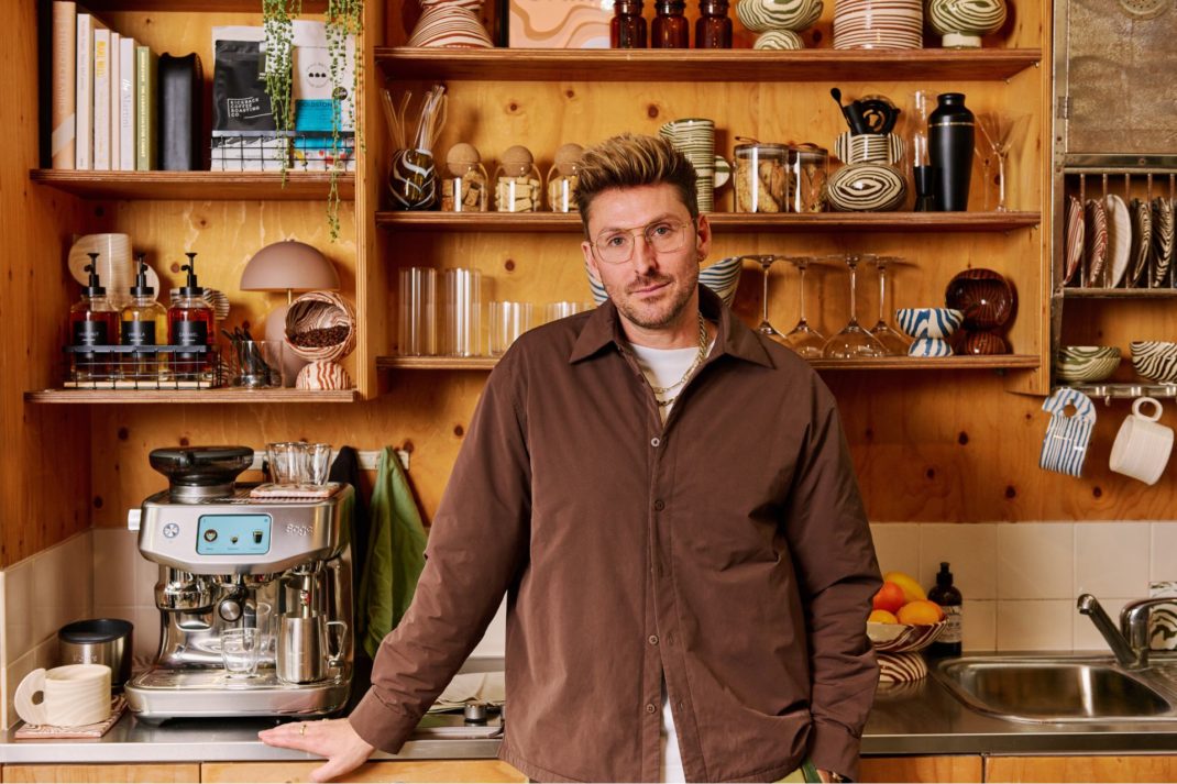 Henry Holland standing in front of a wooden shelf of coffee mugs and a Sage espresso machine