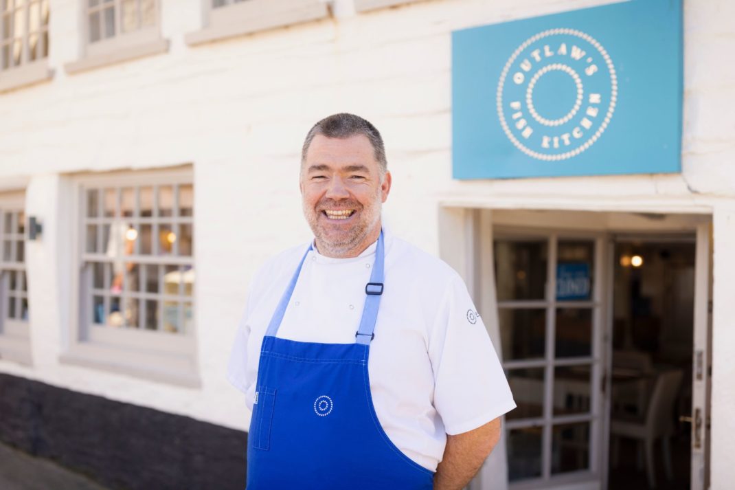 Nathan Outalw standing outside his coastal restaurant wearing a blue apron