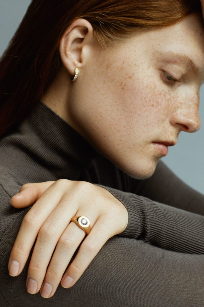 Woman in grey jumperearing a gold Skydiamond signet ring and gold hoop earing