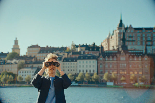 A woman looks into the camera through binoculars. Behind her is water and houses in the centre of Stockholm.