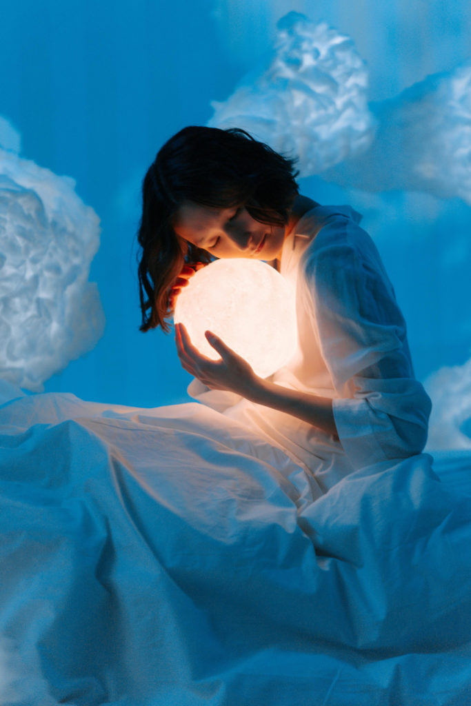 Woman hugging glowing orb in cloudy space with bedding | sleep chronotype