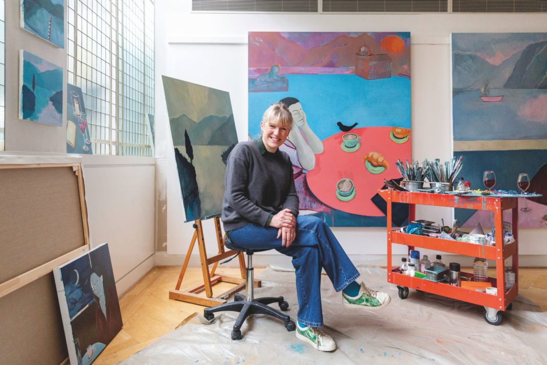 Nancy Cadogan sitting in her studio, with paintings in the background.