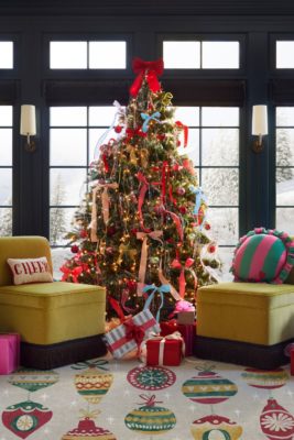 Christmas tree adorned with multicoloured bows, standing on a bauble-patterned rug with two acid yellow armchairs