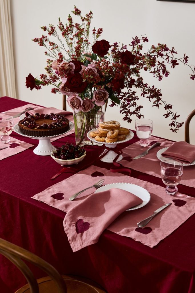 Tablescape with dark red tablecloth, pink placemats with rewd hearts, matching pink napkins, and a vase with a pink bouquet.