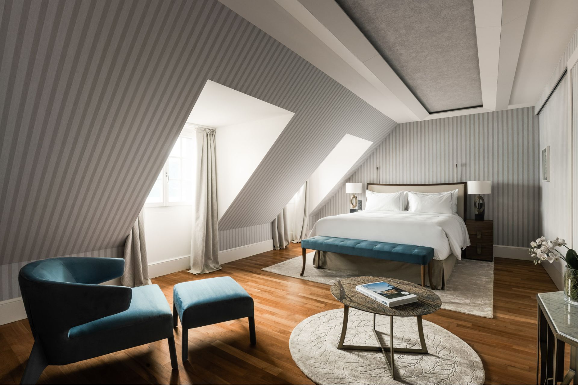 Hotel bedroom with grey striped wallpaper, wooden floors and a cream rug with blue armchairs