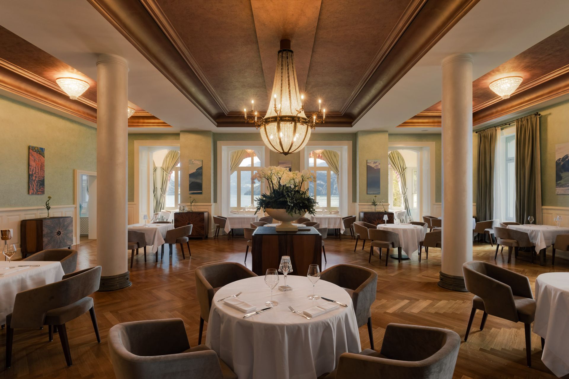 Dining room with tall ceilings, parquet floors, dark grey armchairs and a central chandelier
