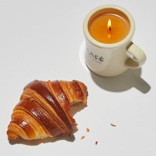 Candle in mug-shaped candle holder with a croissant beside it
