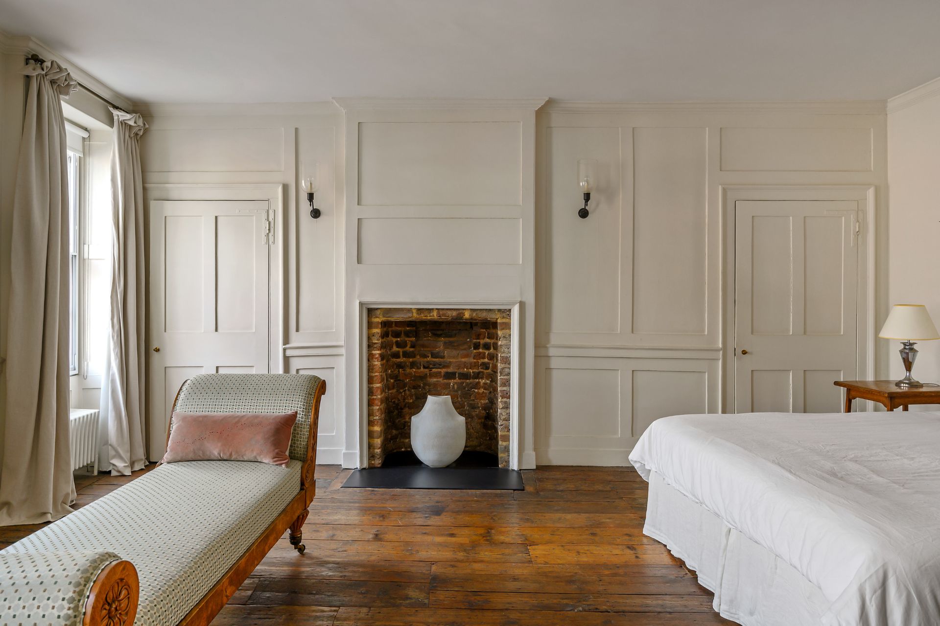 Bedroom with white panelled walls, wood flooring and a brick chimney piece.