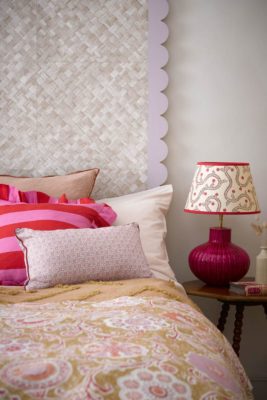 Bed with scalloped white headboard, a yellow bedspread, pink and red striped cushions and a red lamp