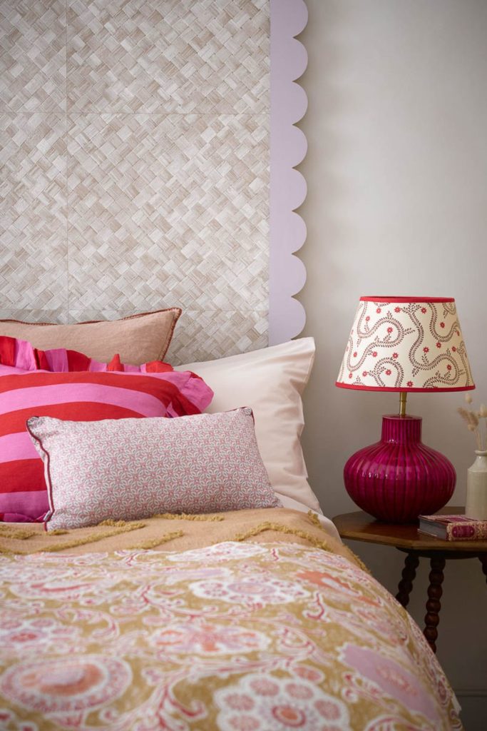 Bed with scalloped white headboard, a yellow bedspread, pink and red striped cushions and a red lamp