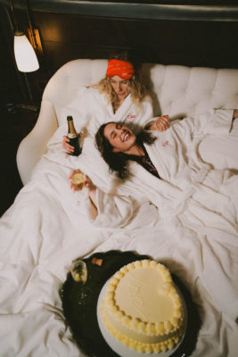 Two women lying in bed in dressing gowns with cake and champagne