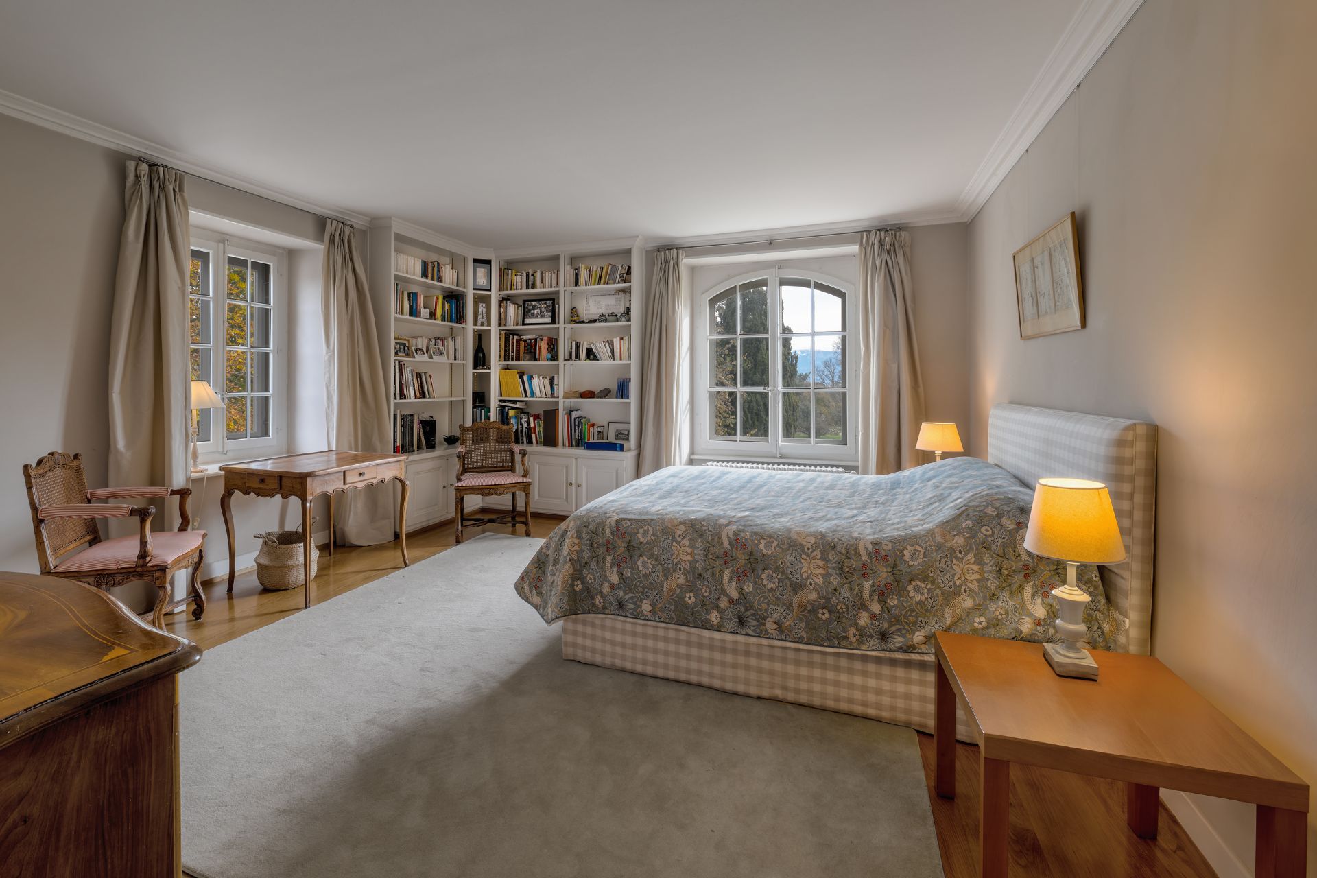 Bedroom with a cream rug, an arched window and a corner bookshelf.