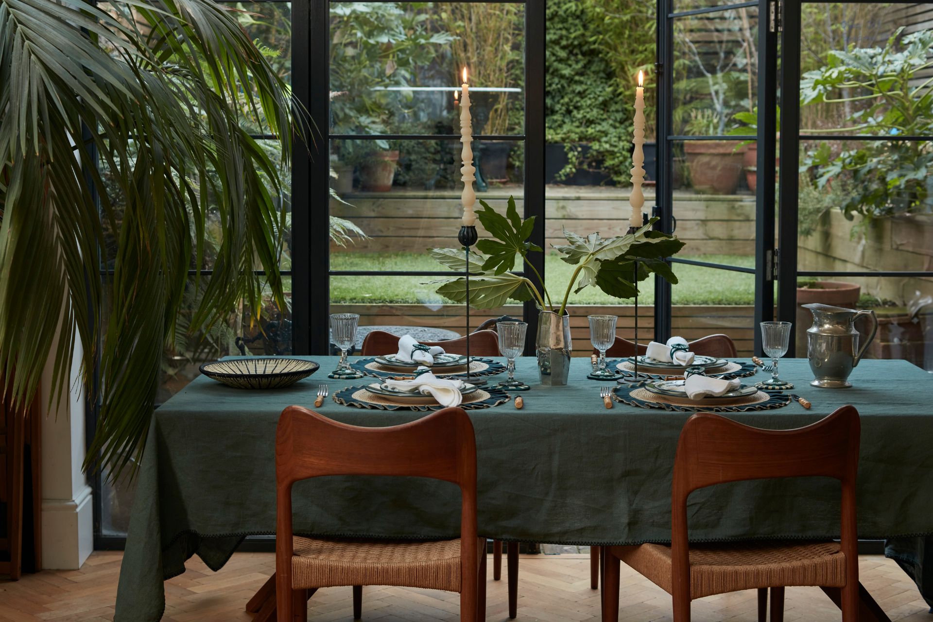 Dining room with Crittall windows, wood and rattan chairs, and a deep green linen tablecloth.