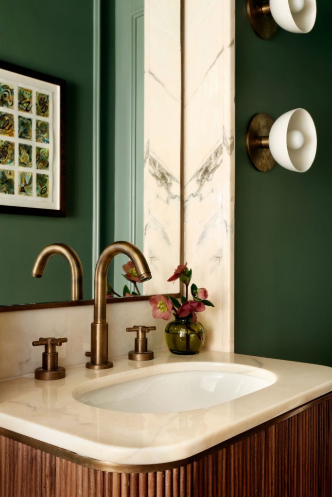 Forest green bathroom with a large mirror, a white marble sink, a fluted wooden cabinet and brass hardware.