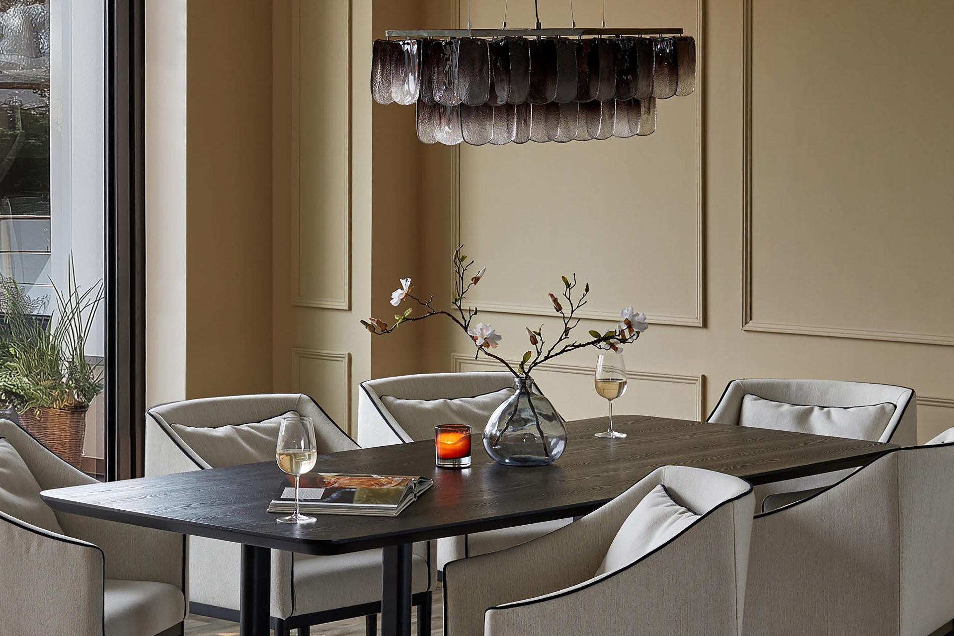 Pale yellow dining room with deep wooden table, cream armchairs and a smoked glass pendant light.