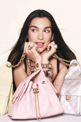 Dua Lipa holding pale pink, yellow and silver Chanel 25 bags, photographed against a pastel orange background.