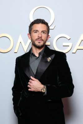Jonathan Bailey wearing a black dinner jacket and grey shirt in front of a pale blue Omega branded backdrop.