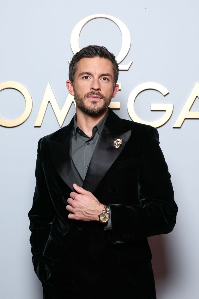Jonathan Bailey wearing a black dinner jacket and grey shirt in front of a pale blue Omega branded backdrop.