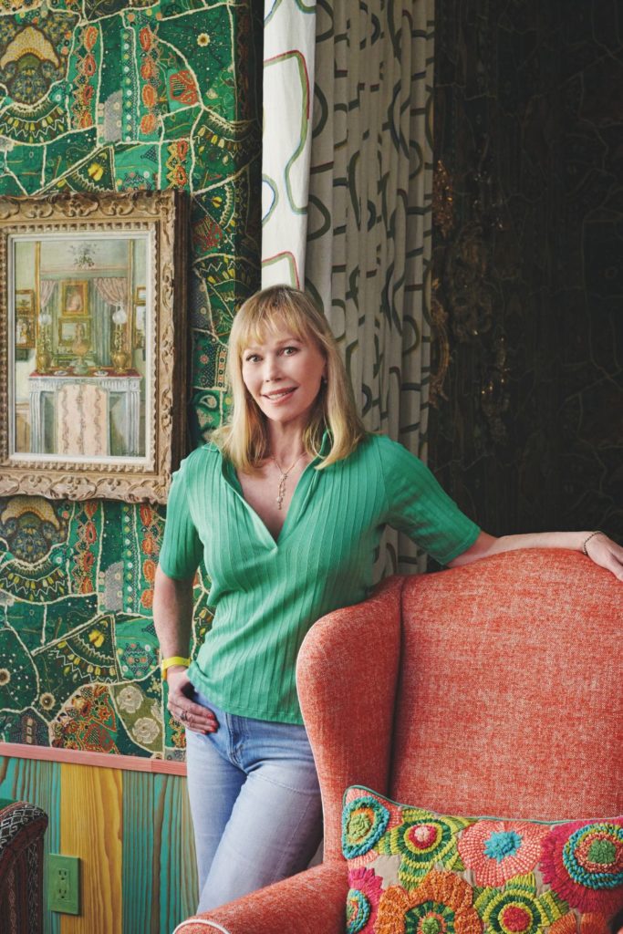 Kit Kemp wearing blue jeans and a green polo neck jumper, standing beside an orange armchair.